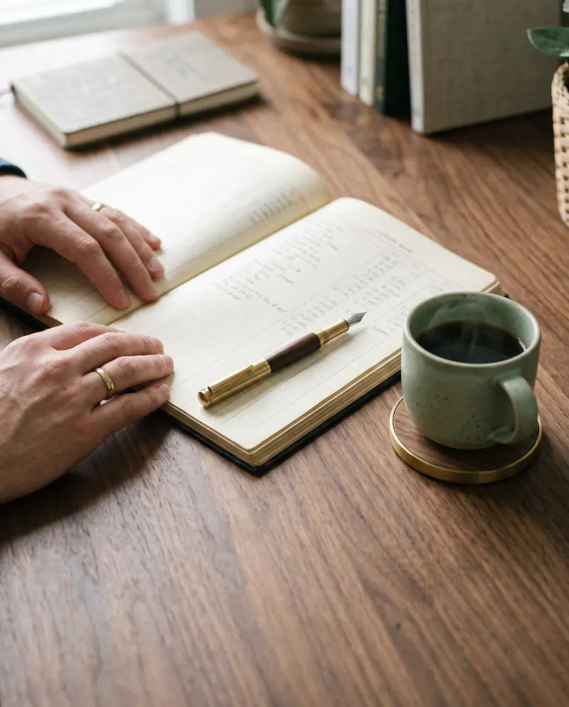 An open ledger, a pen and a ceramic coffee cup on a walnut desk in soft morning light