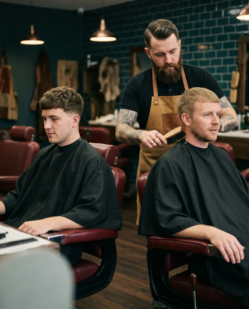 Two men side by side in barber chairs with the same short haircut, full and wavy on one, flatter and finer on the other