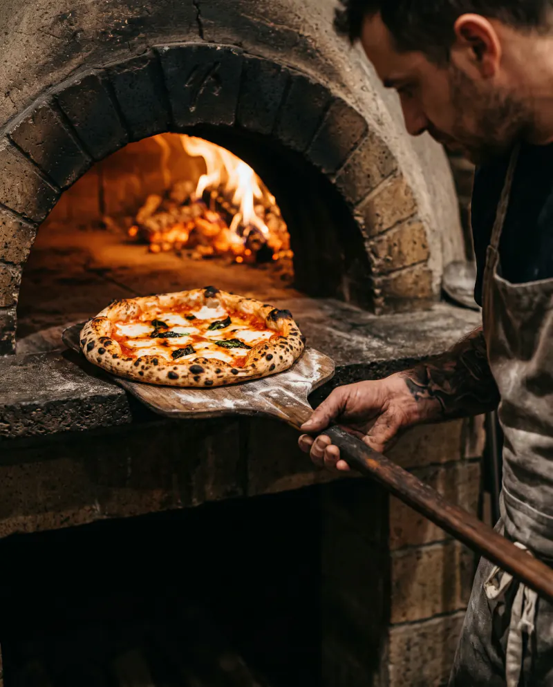 A blistered Neapolitan pizza coming out of a glowing wood-fired oven on a wooden peel